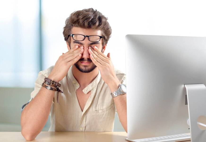 Imagem de um homem esfregando os olhos com as mãos, na frente de um computador.